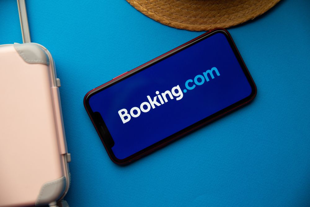 Booking.com logo is shown on phone beside pink suitcase with blue background