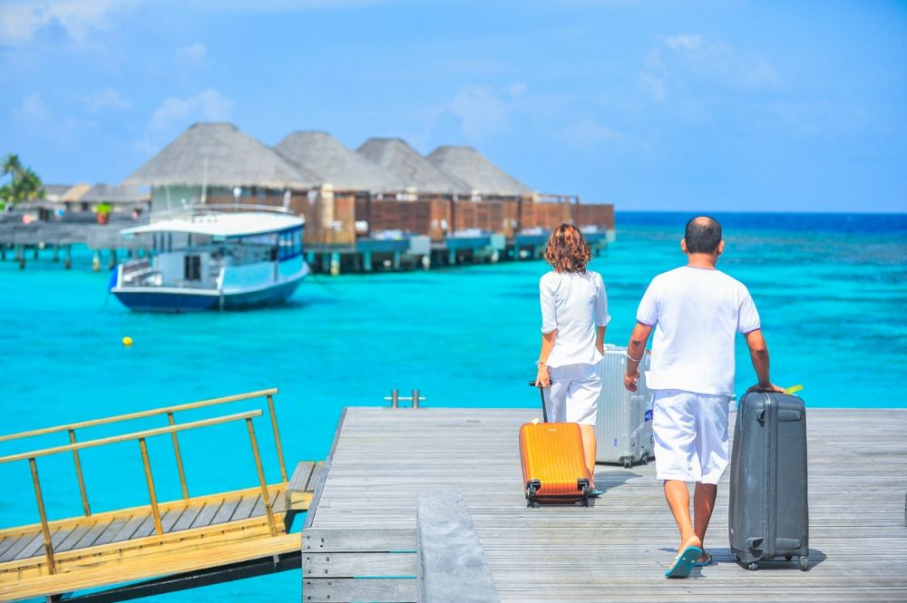 a couple wearing white with suitcases facing floating resort rooms in beach