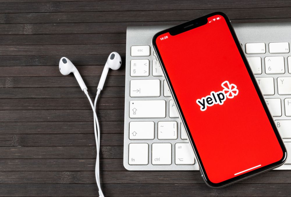 Yelp logo seen on phone on top of an Apple Mac keyboard and Apple earphones set on wooden table